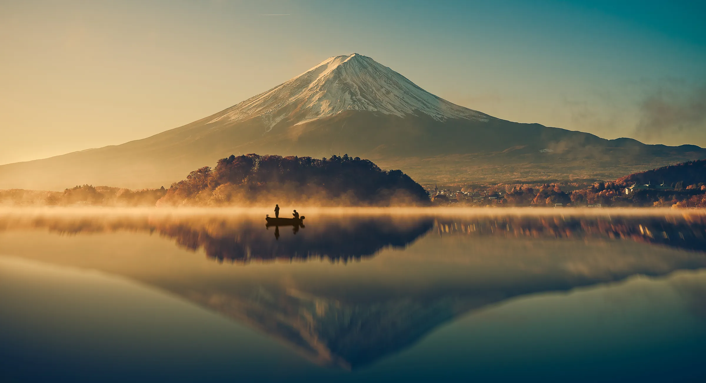 Two people in a small boat on a still, misty lake at sunrise, with Mount Fuji reflected in the water
