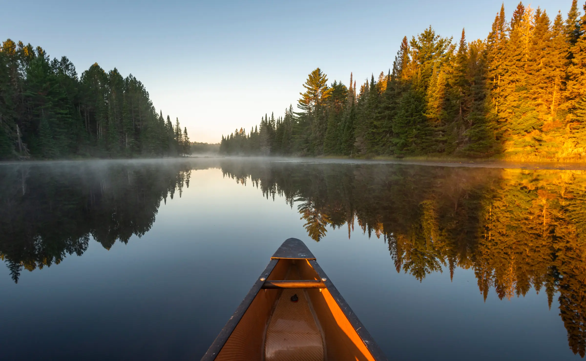The bow of an orange canoe on a still lake at sunrise, pointing down a channel between pine forests
