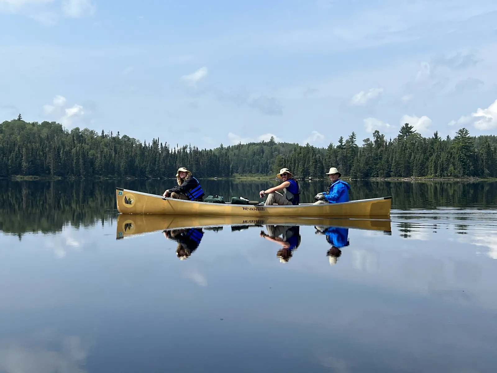 Three paddlers in a canoe on a glassy lake, reflected in the water, with Bill in the stern