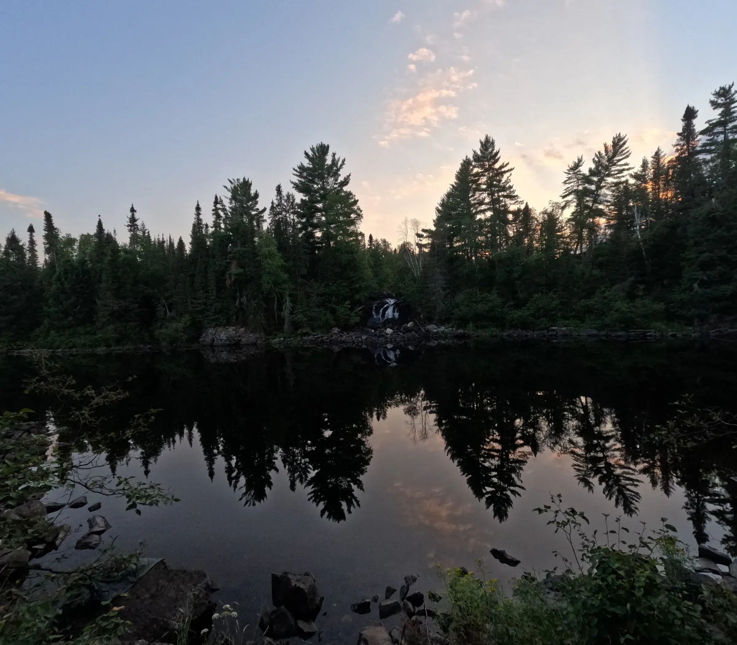 A small waterfall spilling into a still lake at dusk, with pines reflected in the water