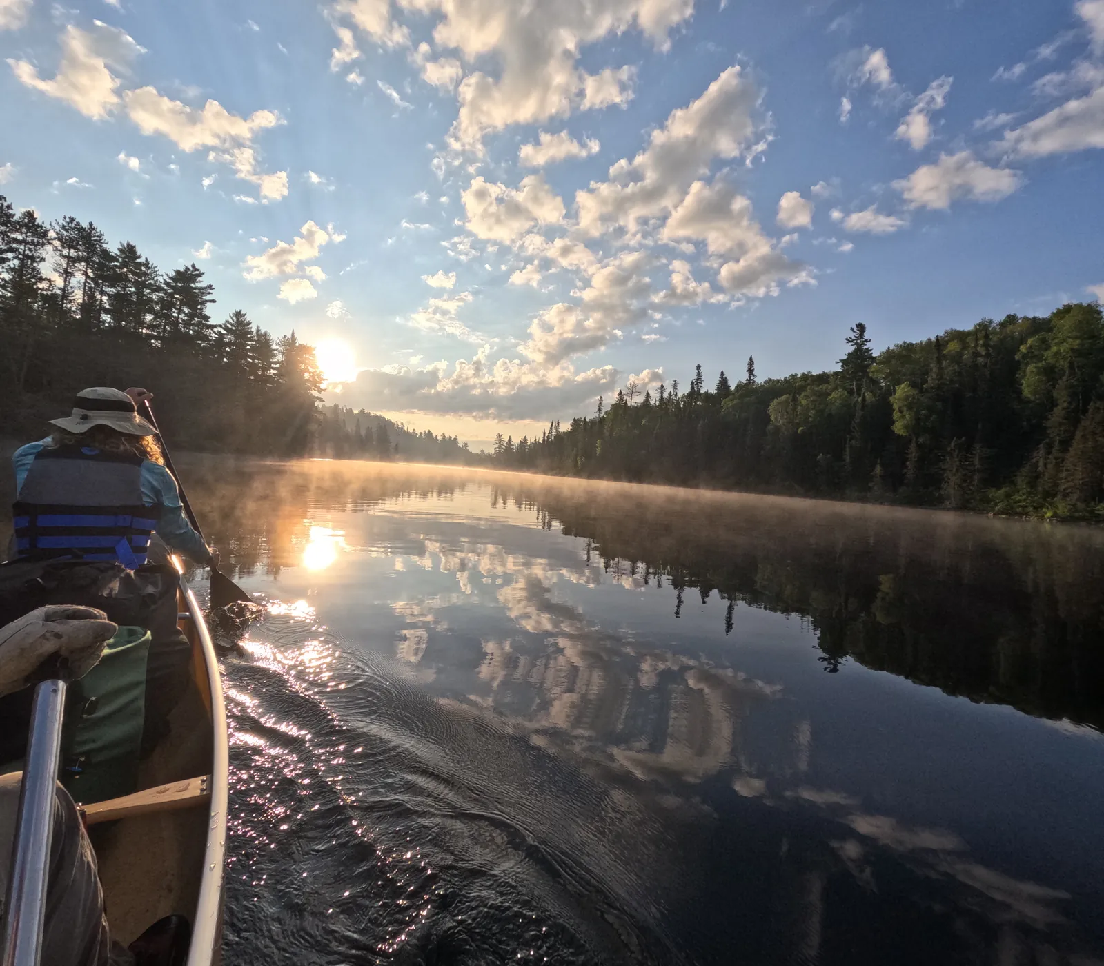 View from a canoe at sunrise as mist lifts off a still lake between pine forests